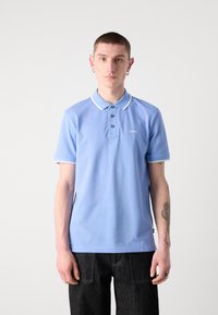 Svetlo modra polo majica z enotno teksturo, trije gumbi na zaponki in beli detajli na ovratniku. Ustrezno kombinirana s temno modrimi kavbojkami, kar daje sproščen videz.