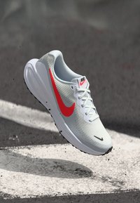Nike bėgimo bateliai šviesiai pilkos spalvos tinklelyje, su ryškiu raudonu swoosh simboliu, balta raišteliu, paminkštintu padu ir tekstūruotais paviršiaus elementais.