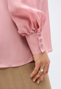 Κοντινό πλάνο ενός χεριού που φορά δαχτυλίδι ασημένιο σε σχήμα λουλουδιού, ακουμπισμένο στο πλάι, με ροζ μπλούζα με φουσκωτά μανίκια και μπεζ φούστα να διακρίνονται.