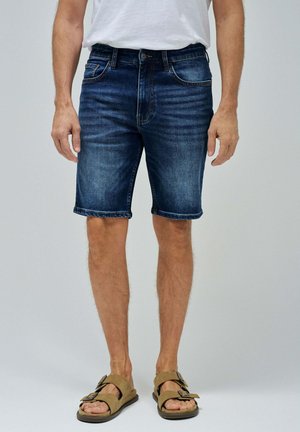 Bermudas de mezclilla en lavado azul oscuro, con cinco bolsillos y corte recto. Estilizadas con un dobladillo enrollado, combinadas con sandalias marrones.