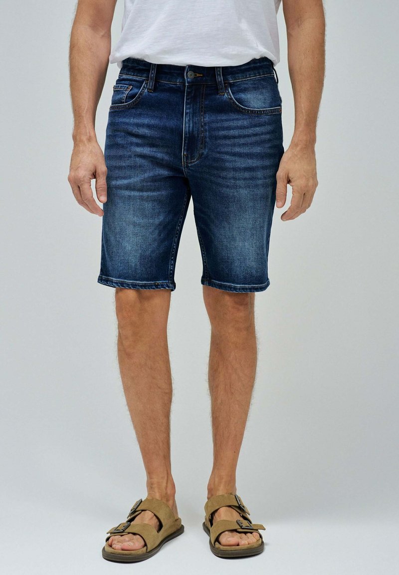 Bermudas de mezclilla en lavado azul oscuro, con cinco bolsillos y corte recto. Estilizadas con un dobladillo enrollado, combinadas con sandalias marrones.