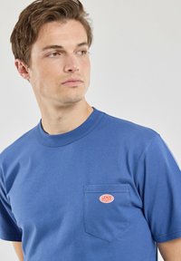 Blaues Baumwoll-T-Shirt mit Rundhalsausschnitt, kurzen Ärmeln und einer linken Brusttasche mit einem rot-weißen Logopatch. Angenehme, glatte Textur.