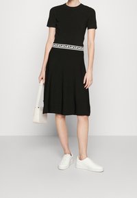 Vestito nero a maniche corte con gonna svasata, dotato di una cintura con logo bianco. Indossato con sneakers bianche e una piccola borsa color crema.