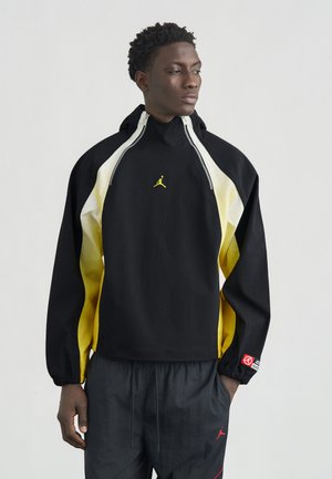 Noor mees, kes kannab musta ja kollast lukuga jope ning musti pükse punase logoga, seisab käed taskutes ja vaatab paremale.