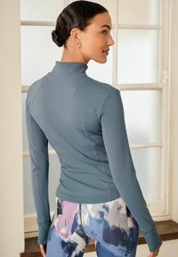 Lyseblå langærmet træningsbluse med høj krave og sømløst design; parret med multikolore, abstrakt-mønstret leggings.