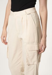 Beige cargobroek met plooien, een zijzak en een knoopsluiting. Gemaakt van een lichtgewicht stof, met een relaxed fit en een gladde textuur.