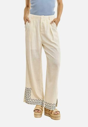 Persona con pantalones de pierna ancha color crema con bordado geométrico negro cerca de los bajos, combinados con sandalias de plataforma tejidas y una camiseta azul de punto acanalado.