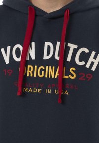 Haut à capuche bleu marine avec cordons rouges. Présente un texte brodé : "VON DUTCH ORIGINALS 1929" en blanc et jaune. Texture de tissu douce.
