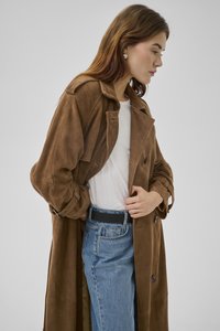 Vrouw met bruin haar, gekleed in een lange bruine trenchcoat, wit shirt, blauwe spijkerbroek en zilveren oorbellen in de vorm van een hart tegen een grijze achtergrond.