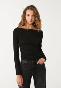 Schwarzes Langarmoberteil mit schulterfreiem Ausschnitt, das einen figurbetonten Schnitt und geraffte Textur aufweist. Kombiniert mit dunkelblauen High-Waist-Jeans.