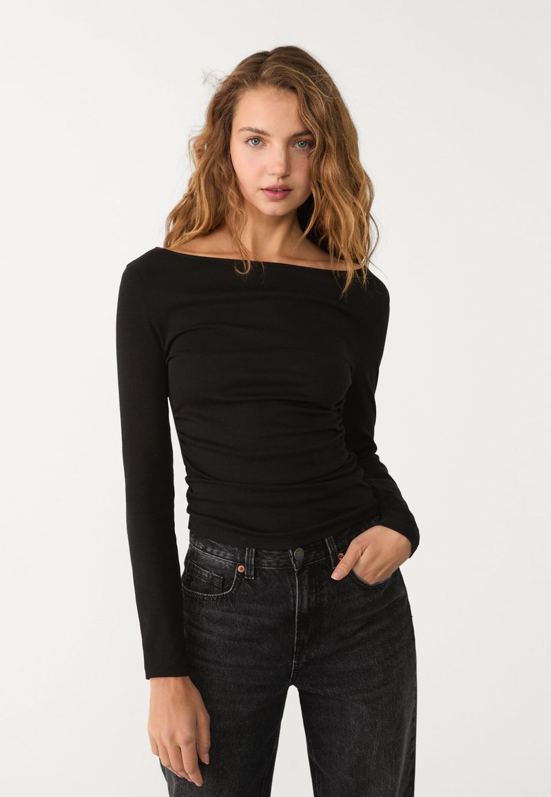 Schwarzes Langarmoberteil mit schulterfreiem Ausschnitt, das einen figurbetonten Schnitt und geraffte Textur aufweist. Kombiniert mit dunkelblauen High-Waist-Jeans.