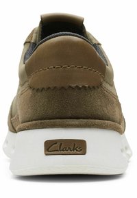Brown suede sneaker with a textured finish, white rubber sole, and a stitched accent. Features a logo tab on the back.