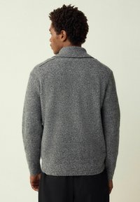 Mies selin päin, yllään harmaa neulepaita ja mustat housut, yksinkertaisen vaalean taustan edessä.