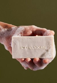 Rechteckige Seife in hellbeige, die in einer Hand gehalten wird, umgeben von weißem Schaum, mit "(MALIN+GOETZ)" auf der Oberfläche geprägt.