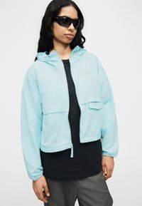 Νεαρός άνθρωπος που φοράει μαύρα γυαλιά ηλίου, ανοιχτό μπλε μπουφάν με φερμουάρ πάνω από μαύρο πουκάμισο και γκρι παντελόνια, στέκεται μπροστά σε απλό λευκό φόντο.