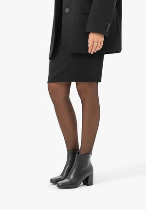 Femme portant des bottines noires à talons carrés, des collants noirs transparents, une jupe noire et un blazer noir, debout sur un fond blanc.