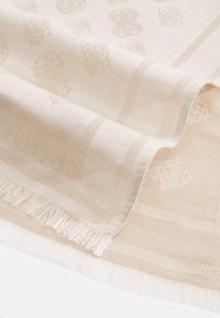 Beige gewebter Stoff mit dezenten Blumenmustern, weicher Textur und fransigen Rändern, sanft auf einer weißen Oberfläche drapiert.