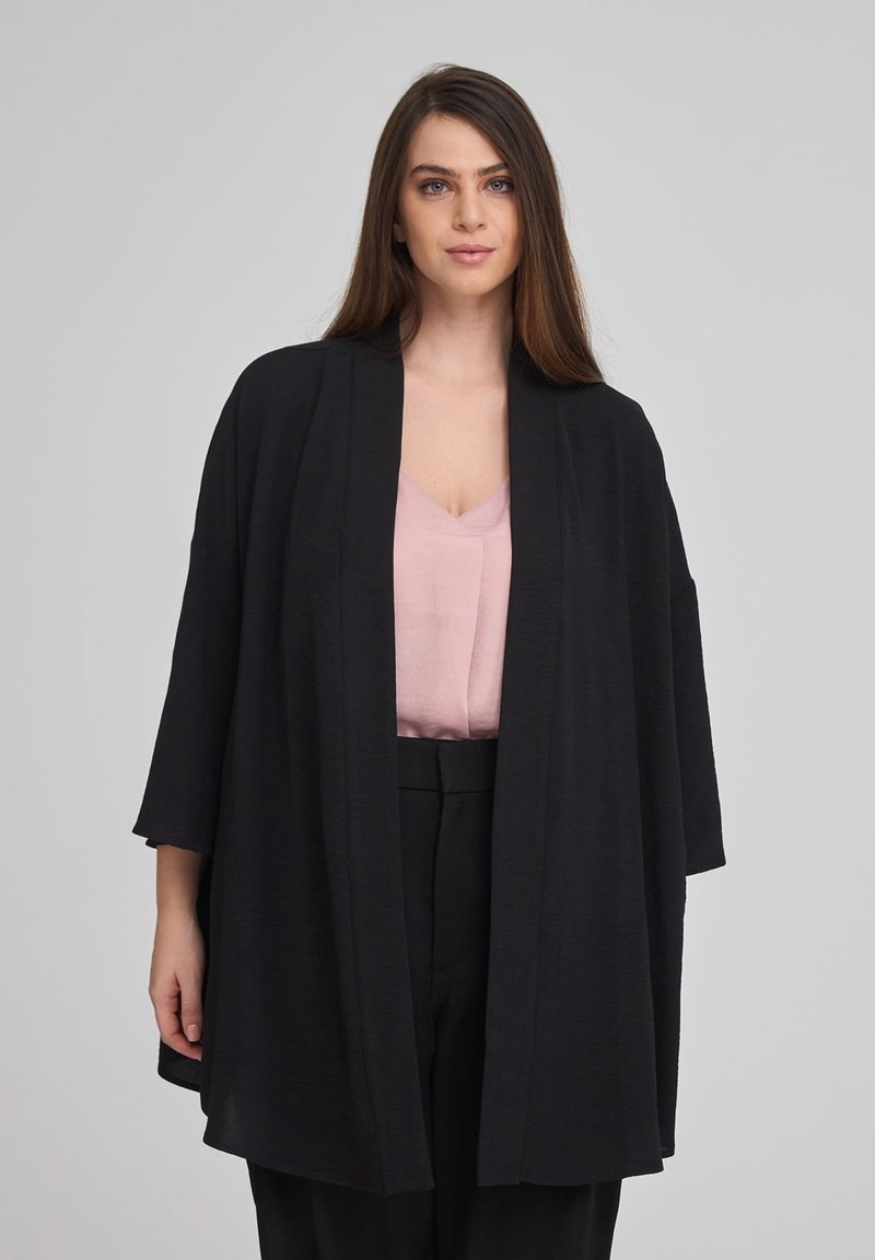 Kimono nero aperto davanti realizzato in tessuto morbido, caratterizzato da maniche ampie e una vestibilità rilassata, indossato sopra una maglietta rosa chiaro e pantaloni neri.