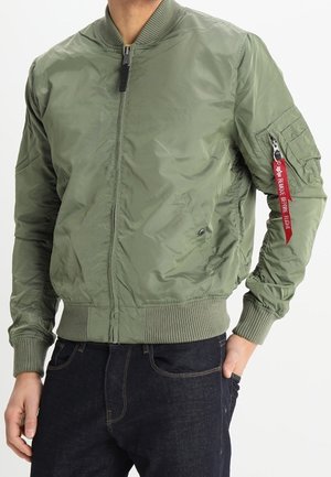 Homme portant un blouson aviateur vert olive avec poignets et ourlet côtelés, étiquette rouge « Remove Before Flight » sur la poche de la manche et jean foncé.