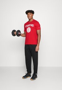 T-shirt rossa in cotone con logo "Stanford", pantaloni sportivi neri e sneakers nere con dettagli marroni, in mano destra tiene un manubrio nero.