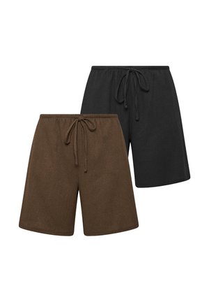 Vrouw met lang donker haar, die een beige mouwloze gebuttonde top draagt, bruine shorts tot aan de knie en bruine sandalen, staand tegen een eenvoudige muur.