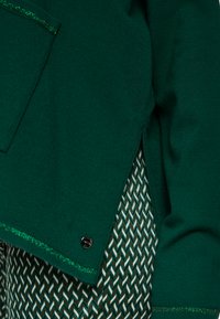 Der grüne Pullover weist ein strukturiertes Material mit einem metallischen Akzent am Saum auf. Ein gemusterter Stoff darunter zeigt ein Fischgrätenmuster.