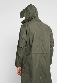 Cappotto impermeabile lungo di colore verde oliva, dotato di cappuccio, vita regolabile con cordino e polsini con bottoni. Tessuto liscio e leggero con finitura opaca.