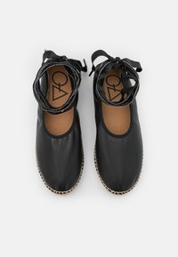 Sapatos planos pretos de couro com uma superfície lisa. Apresentam bico redondo, fitas no tornozelo e solas texturizadas com bordas trançadas.