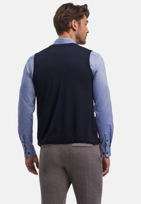 Gilet di maglia blu marina sopra una camicia azzurra chiara, con un taglio classico. L'outfit è completato da pantaloni grigi con una texture liscia.