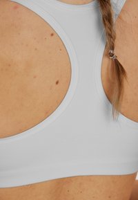 Ljust grå sportbh med scoop-back design, tillverkad av mjukt material och prydd med prägla "ENDURANCE"-logotyp, synlig sömnad och kurvad siluett.