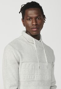 Hellgrauer Hoodie mit strukturiertem Muster, ausgestattet mit einem hohen Kragen und Kordelzug. Prägiertes Logo auf der Brust, weicher Stoff.