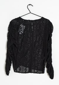 Blusa di pizzo nero con maniche a palloncino, motivo floreale strutturato, scollatura rotonda e cuciture verticali, appesa a un gancio nero.