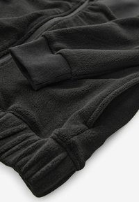 Schwarze Fleece-Jacke mit gerippten Bündchen, ausgestattet mit einem Reißverschluss und weichem Material. Sichtbare Nähte und geraffte Taillendetails.