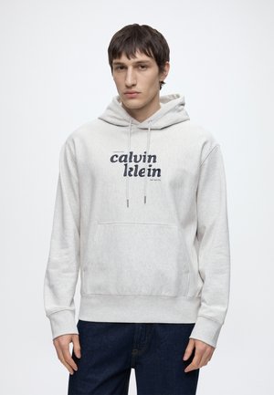 Νέος άνδρας που φοράει ανοιχτό γκρι φούτερ Calvin Klein με τσέπη μπροστά και σκούρο μπλε τζιν, στέκεται μπροστά σε ένα απλό λευκό φόντο.