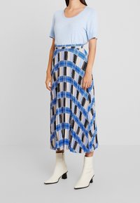 Top azzurro chiaro a maniche corte abbinato a una gonna plissettata con un motivo geometrico nero e blu, e stivali bianchi alla caviglia con tacco a blocco.