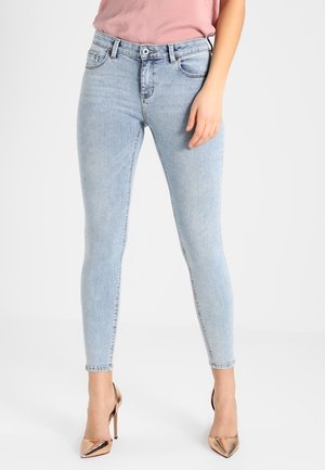 Vrouw draagt lichtblauwe skinny jeans en metallic roségouden hoge hakken, staand tegen een effen achtergrond.