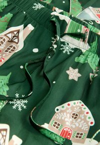 Green fabric shorts featuring a festive design with white decorative snowflakes, gingerbread houses, and holiday trees. Elastic waistband detail.
