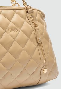 Beige gewatteerde handtas van soepel faux leer. Heeft gouden hardware, een ritssluiting en een subtiel gerepousseerd logo.