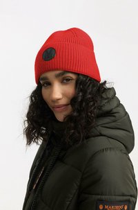 Berretto di lana rosso con una texture a coste e una toppa rotonda nera. Abbinato a un giubbotto puffer verde scuro con lacci regolabili.