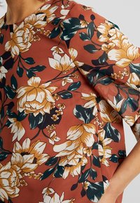 Blusa floreale con grandi fiori bianchi e gialli su uno sfondo ruggine. Maniche corte e leggermente bombate, con una texture liscia.