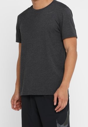 Mann trägt ein schlichtes dunkelgraues kurzärmliges T-Shirt und schwarze Shorts und steht vor einem schlichten Hintergrund.