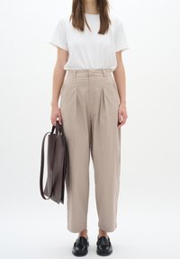 Béžové kalhoty s vysokým pasem a sklady, spárované s bílým tričkem. Černé mokasíny a hnědá kožená taška dotvářejí celý outfit.