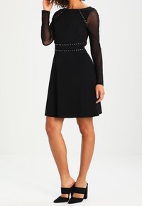 Schwarzes Kleid mit langen transparenten Ärmeln, tailliertem Oberteil und schwingendem Rock. Mit silbernen Nieten entlang des Ausschnitts und der Taille. Getragen mit schwarzen Absatzschuhen.