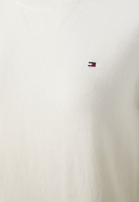 Bílý svetr s kulatým výstřihem, vyrobený z měkké pleteniny. Má na hrudi malé vyšívané logo v námořnické modři, červené a bílé barvě.