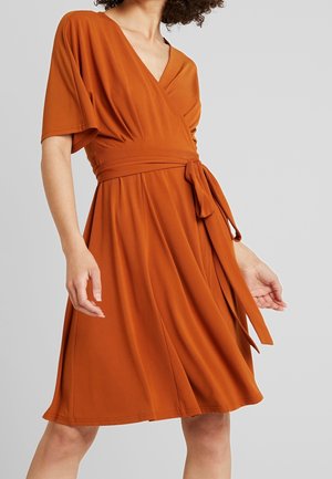 Žena v oranžově rezavých zavinovacích šatech ke kolenům s krátkými rukávy, véčkovým výstřihem a zavázaným páskem v pase, na jednobarevném šedém pozadí.