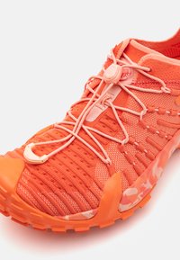 Oransje treningssko med en teksturert nettingoverdel, elastisk snøresystem og polstret krage; har en såle med camo-mønster og slitesterk gummitåhette.