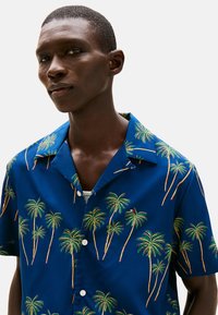 Camisa de manga corta con cuello en tela azul que presenta un vibrante patrón de palmeras en verde y amarillo, con botones blancos en la parte delantera.