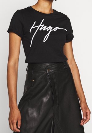 Persona che indossa una t-shirt nera con il testo bianco "Hugo" e una gonna di pelle nera a vita alta con bottoni davanti e dettagli delle cuciture.