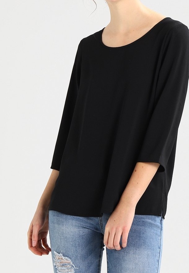 Femme portant un haut noir ample à manches trois-quarts associé à un jean bleu clair déchiré, sur un fond uni.