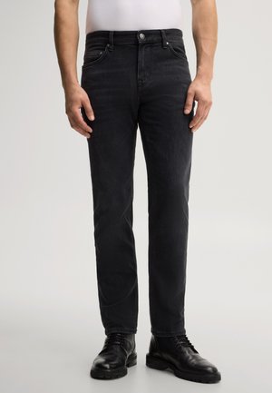 Homme portant un jean noir à jambes droites, des chaussures en cuir noires et une chemise blanche, debout sur un fond gris clair.